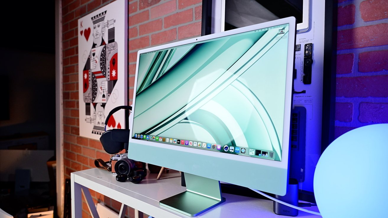 A modern computer with a green-themed display sits on a desk, surrounded by a camera, headphones, and colorful wall art behind.