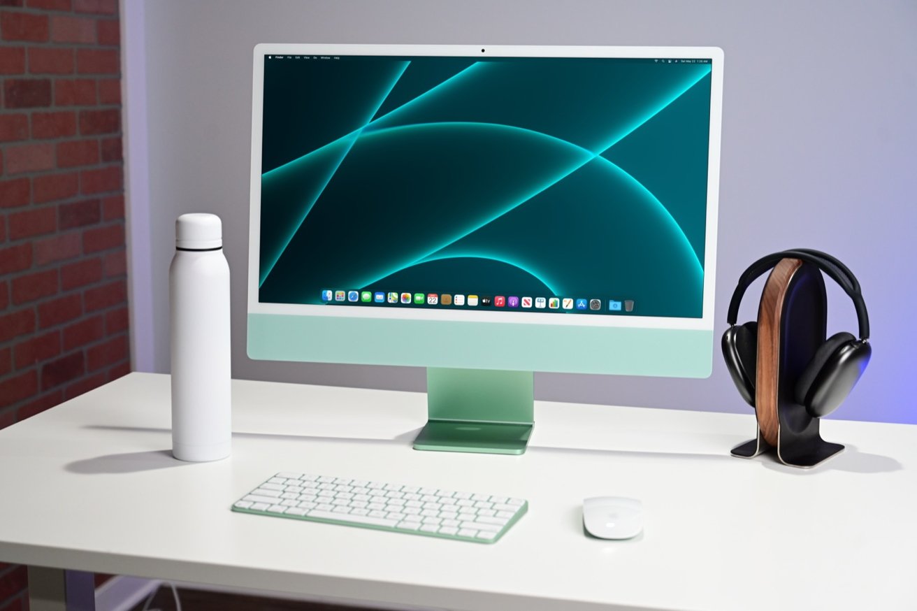 A computer monitor on a desk with a keyboard, mouse, white bottle, and black headphones on a wooden stand.