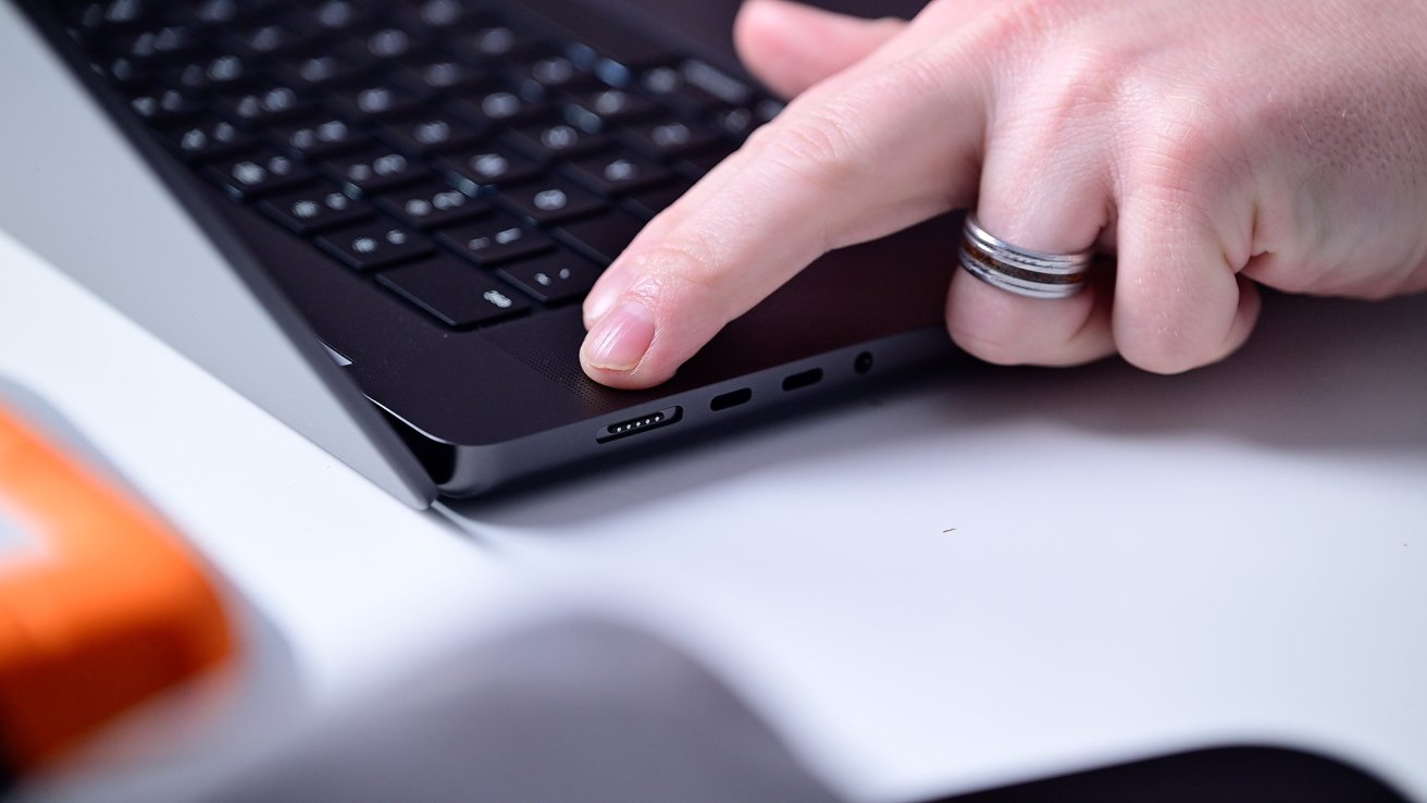 A hand with a ring touches a black laptop near the keyboard and charging port on a white table.